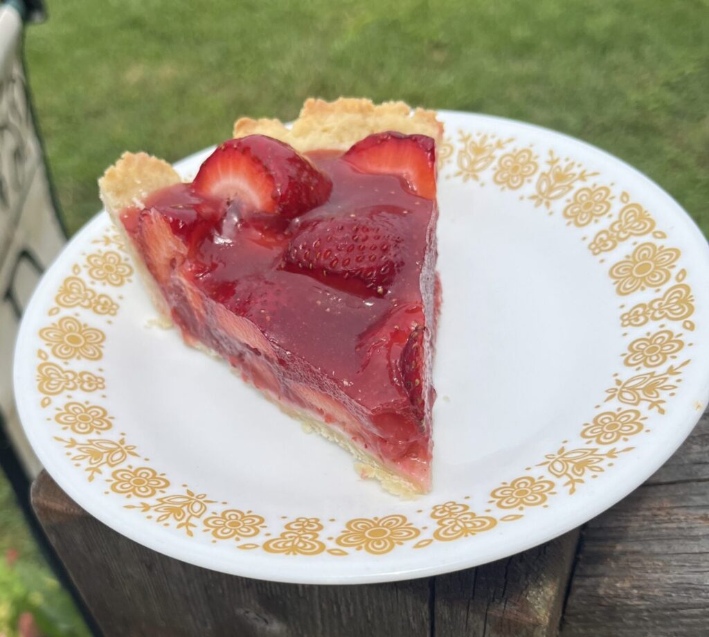 Slice of strawberry pie on a small dessert plate with a background of flowers and grass.